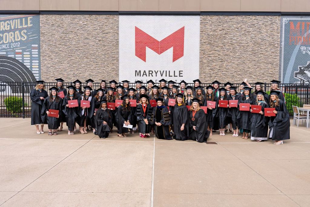 class of graduating students, diplomas in hand, in front of a brick wall with the Maryville logo.